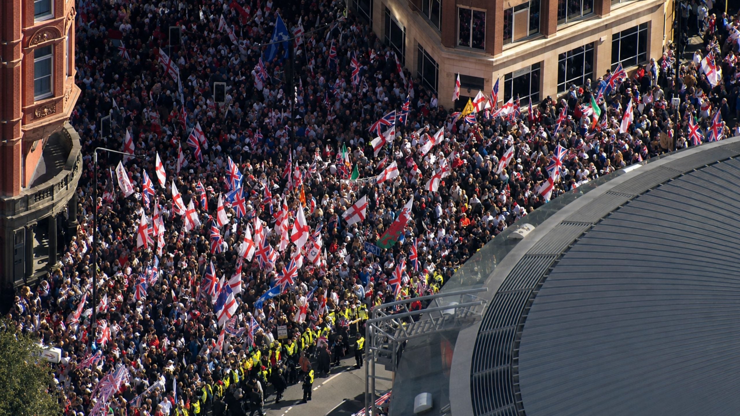 Large crowded street protest with many people waving Union Jack and St George flags between city buildings, police in high-visibility vests lining the road.